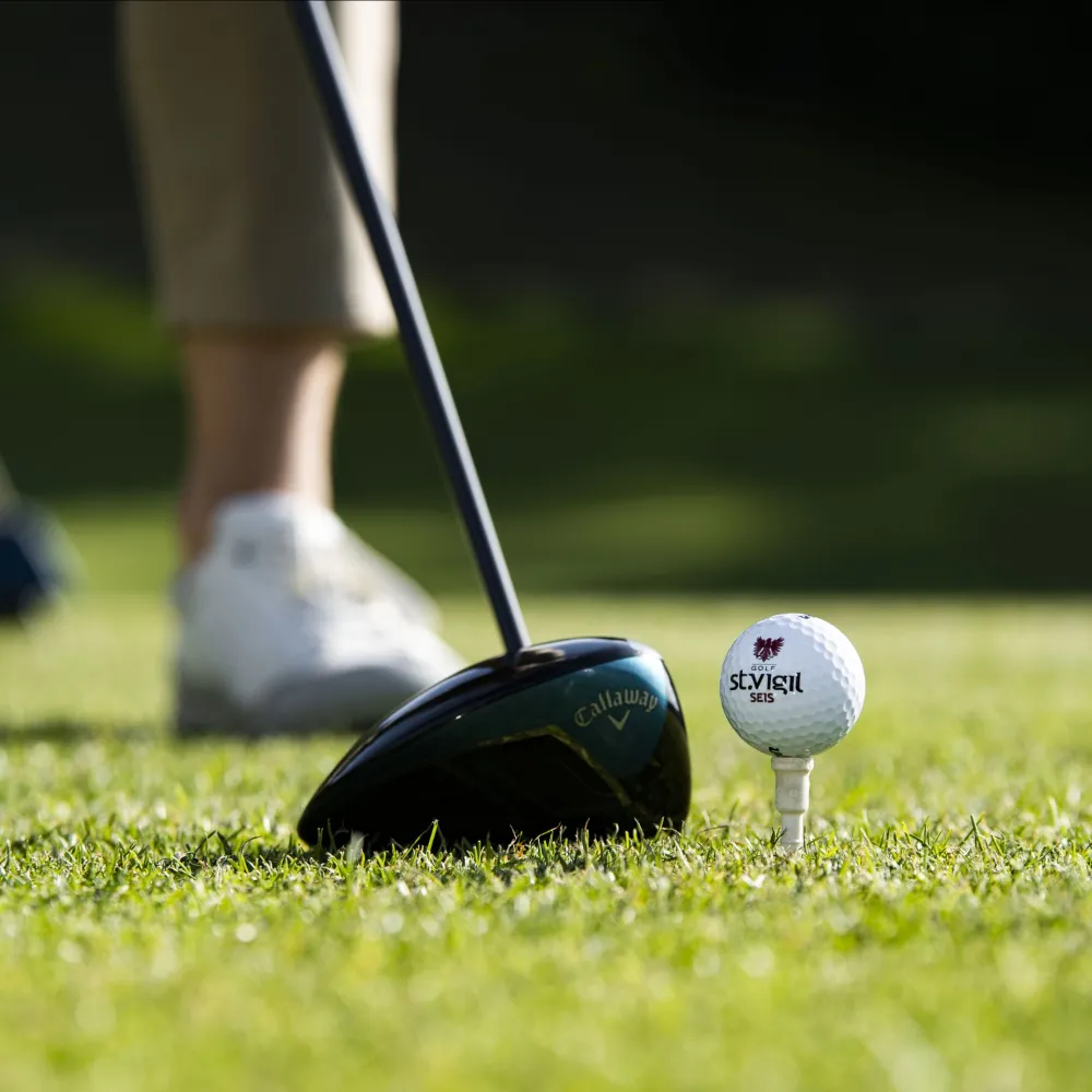 Golf ball with the St. Vigil logo on the tee beside a driver at the golf course in Castelrotto