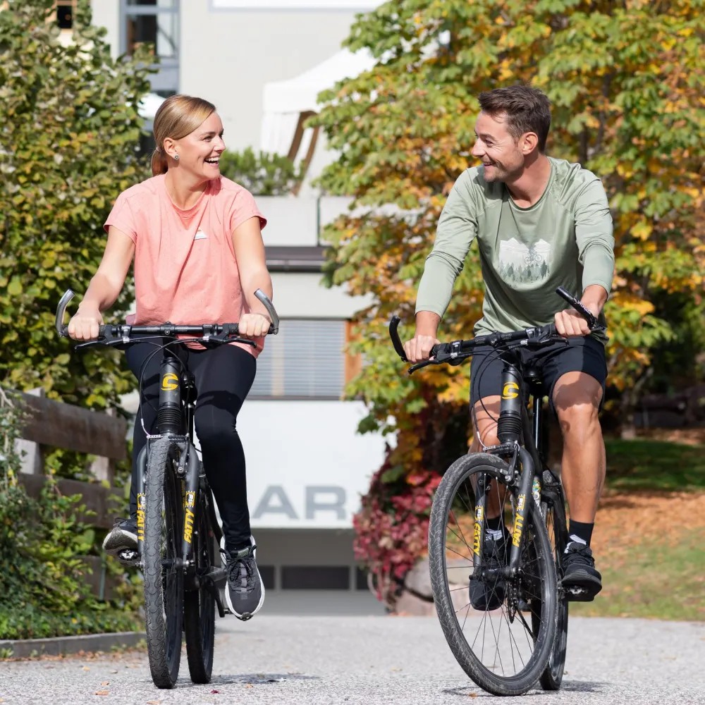 Smiling couple on bikes in front of the Wellness Hotel Alpenflora on a sunny day