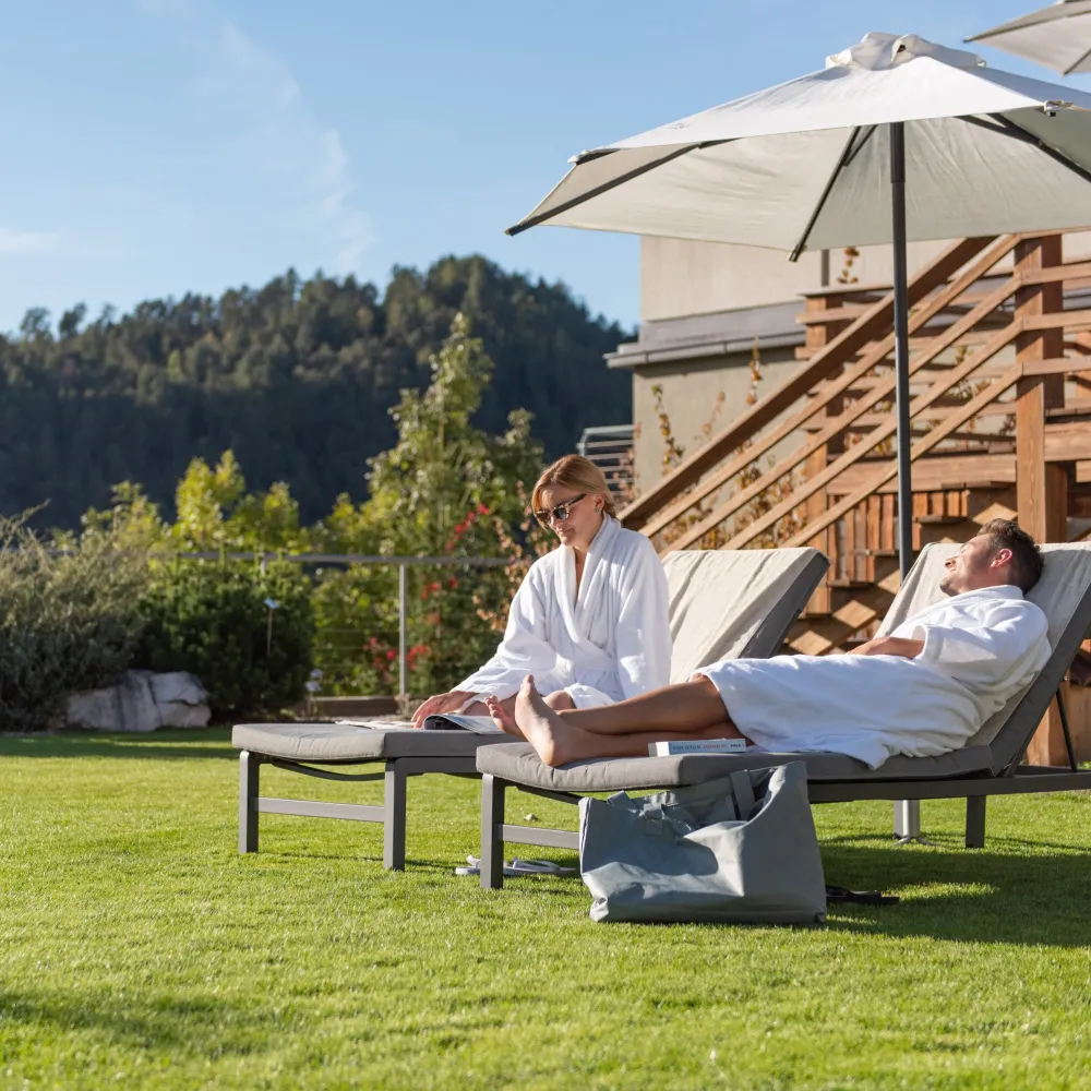 Paar in weißen Bademänteln entspannt auf Liegen unter einem Sonnenschirm im Garten des Wellnesshotels Alpenflora