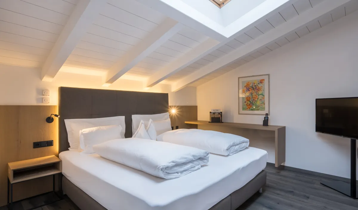 Zona notte dell'Attico Suite nel Wellnesshotel Alpenflora con letto matrimoniale, lucernario e soffitto con travi bianche.