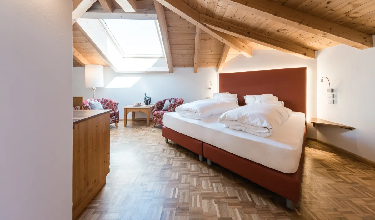 Doppelzimmer Attico im Wellnesshotel Alpenflora mit Doppelbett, Dachfenster, Holzbalkendecke und zwei gemusterten Sesseln.