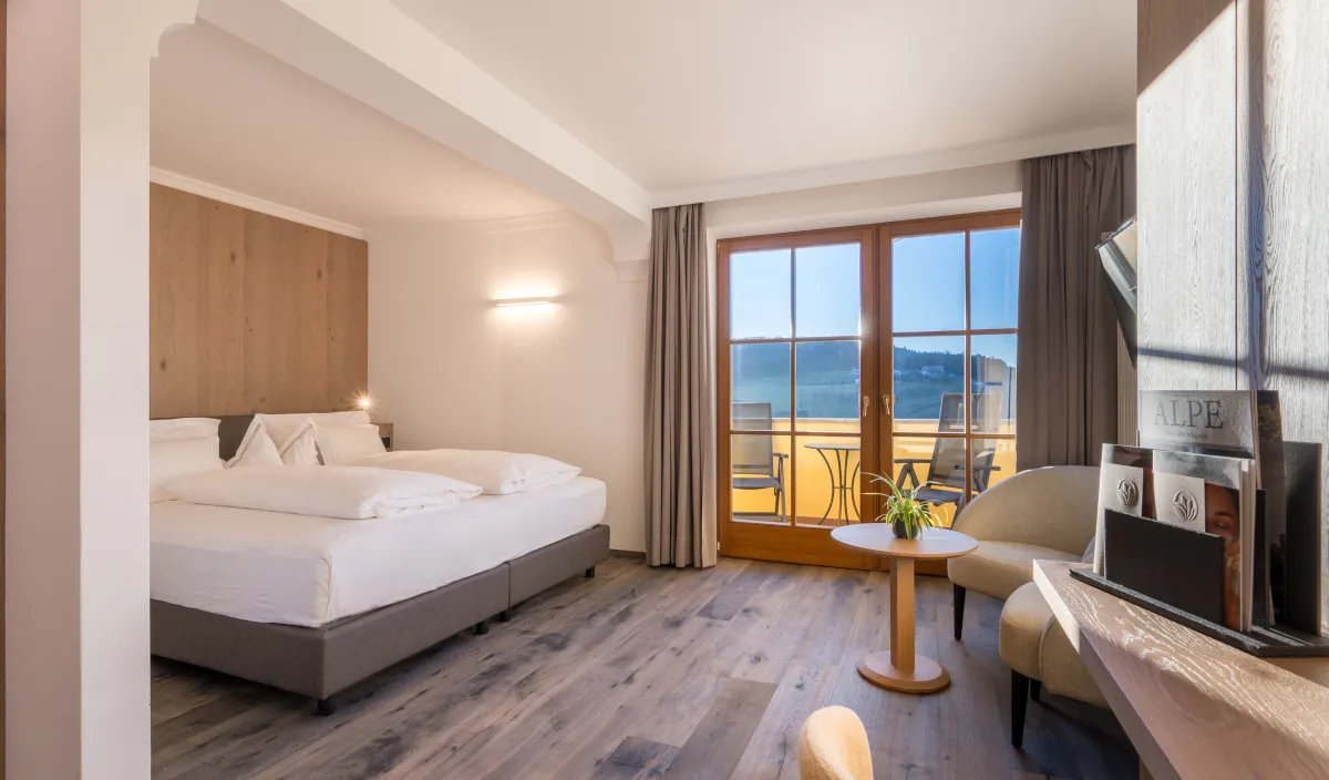 Doppelzimmer Panorama Superior im Wellnesshotel Alpenflora mit Doppelbett, Sessel und Balkontür mit Blick ins Grüne.