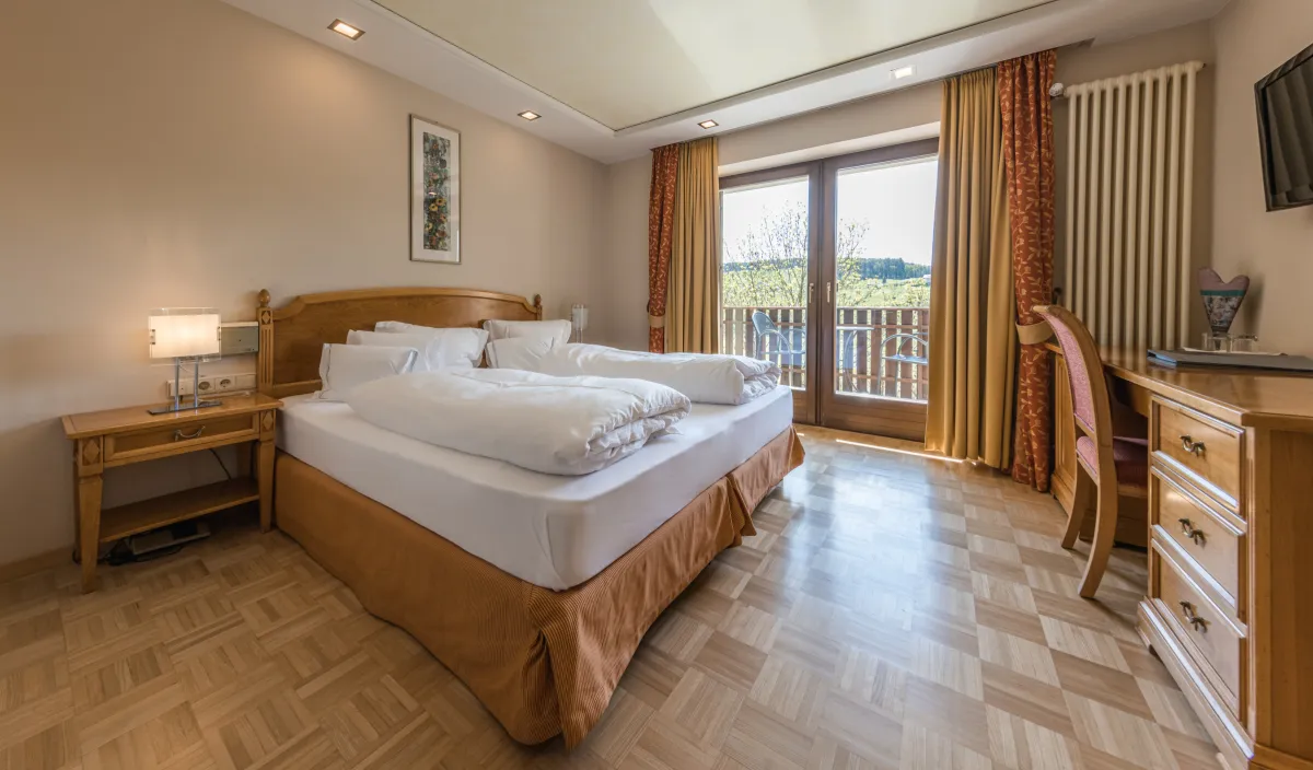 Doppelzimmer Sun im Wellnesshotel Alpenflora mit Doppelbett, Nachttisch, Schreibtisch und Balkontür mit Blick ins Grüne.