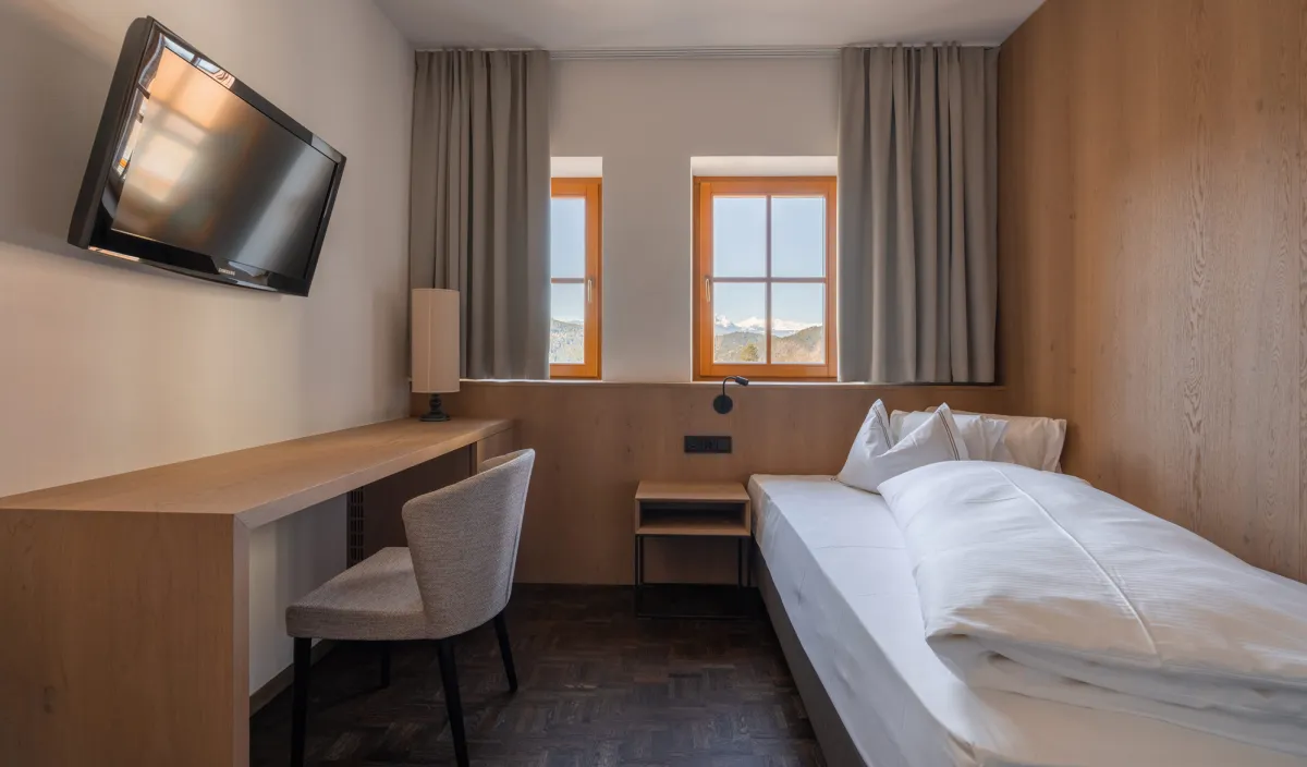 Camera singola Single Stay nel Wellnesshotel Alpenflora con letto, scrivania, televisore e finestre con vista sulle montagne.