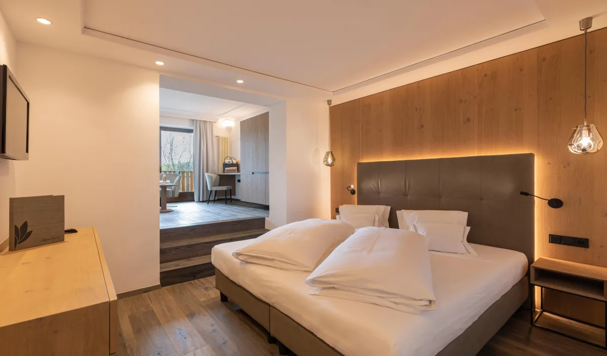 Zona notte della Sun Suite nel Wellnesshotel Alpenflora con letto matrimoniale, rivestimento in legno e passaggio al soggiorno.