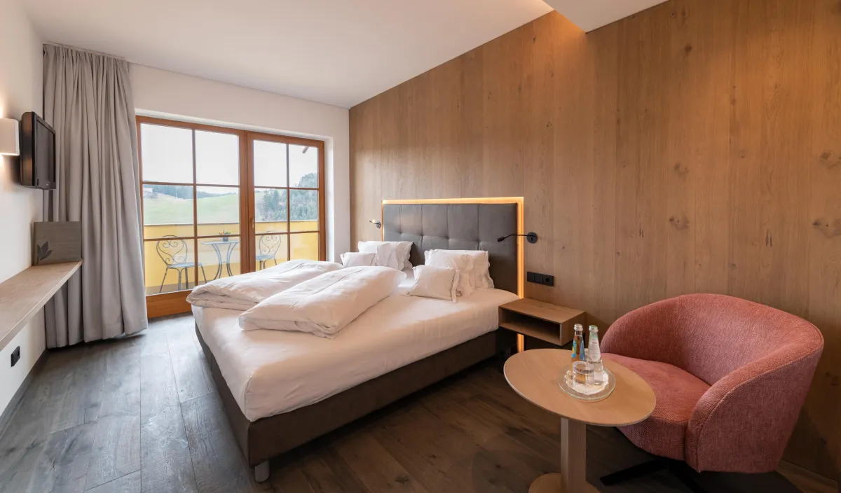 Letto matrimoniale con testiera imbottita, rivestimento in legno, poltrona rosa e porta del balcone con vista sul verde.