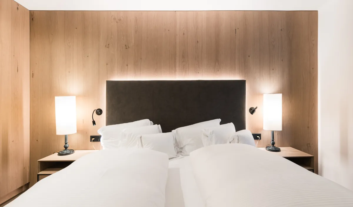 Doppelbett mit anthrazitfarbenem Kopfteil, indirekter Beleuchtung und Holzwandverkleidung im Zimmer Classic.