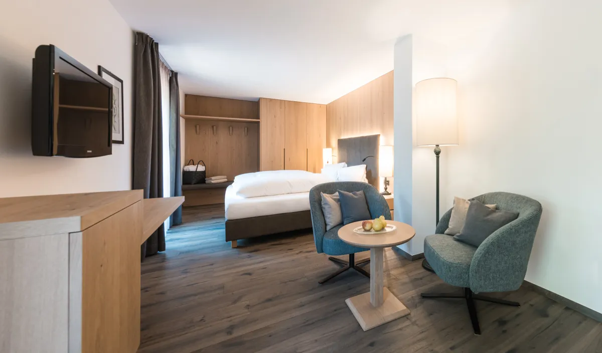 Doppelzimmer Classic mit zwei Sesseln, Beistelltisch, Stehleuchte, Fernseher und Doppelbett im Hintergrund.