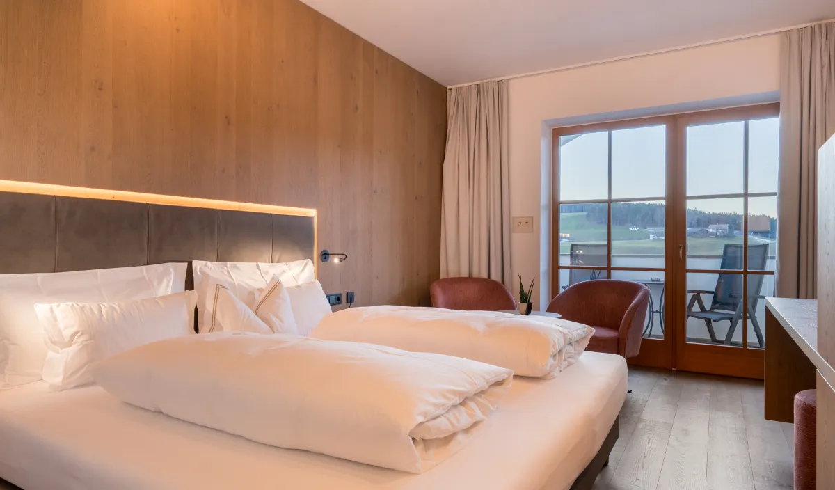Letto matrimoniale con rivestimento in legno, poltrona bordeaux e porta del balcone con vista su prati e colline.