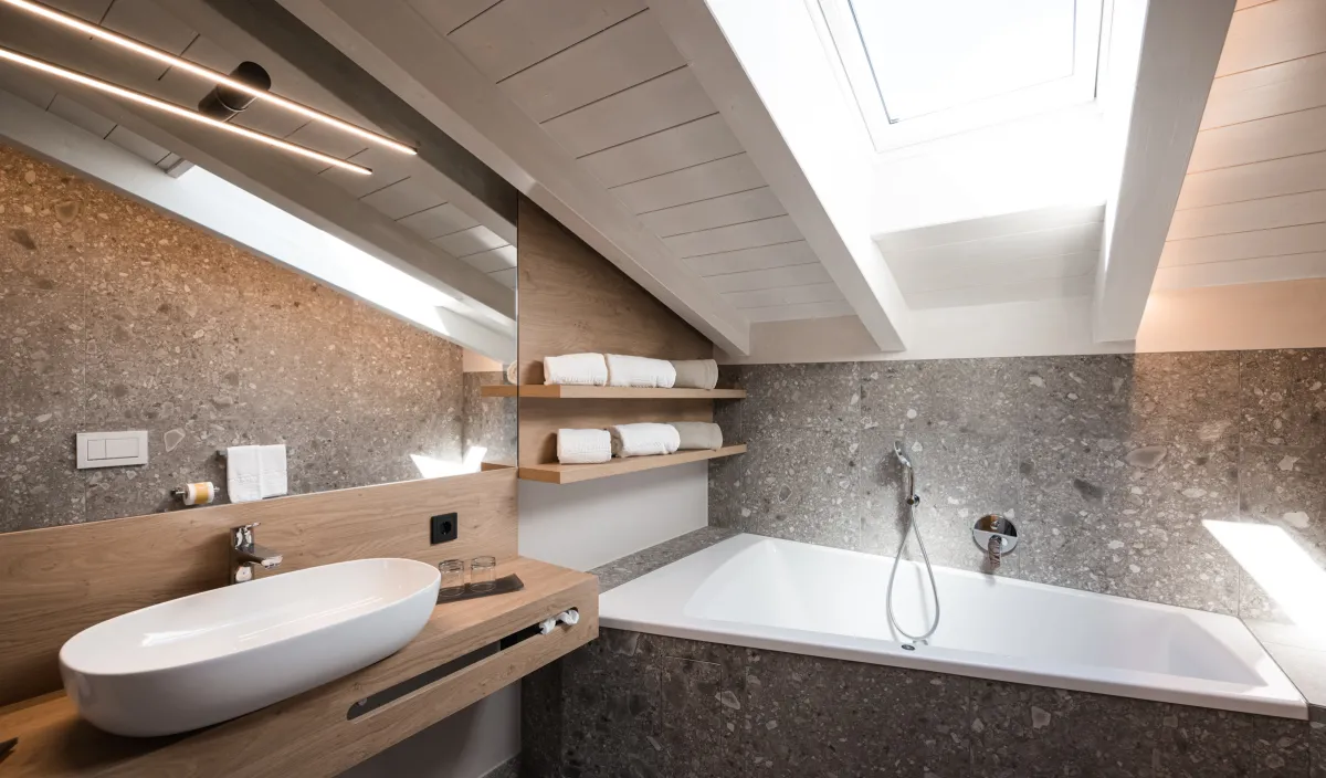 Badewanne unter der Dachschräge mit Dachfenster, Holzwaschtisch, Naturstein-Terrazzofliesen und Handtuchregalen.