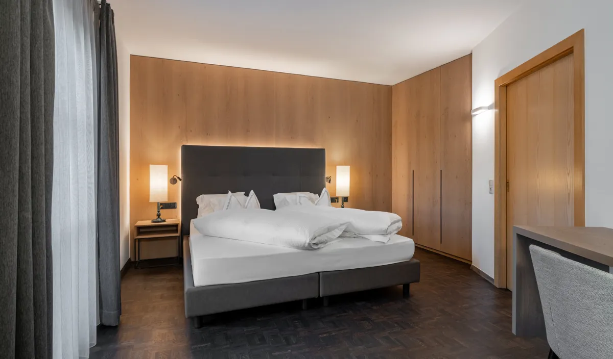 Doppelbett mit anthrazitfarbenem Kopfteil, Holzwandverkleidung, Einbauschrank und Schreibtisch im Zimmer Classic.