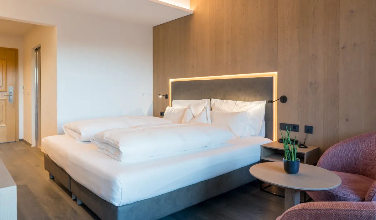 Letto matrimoniale con nicchia illuminata sulla testiera, rivestimento in legno, comodino e poltrona rosa.