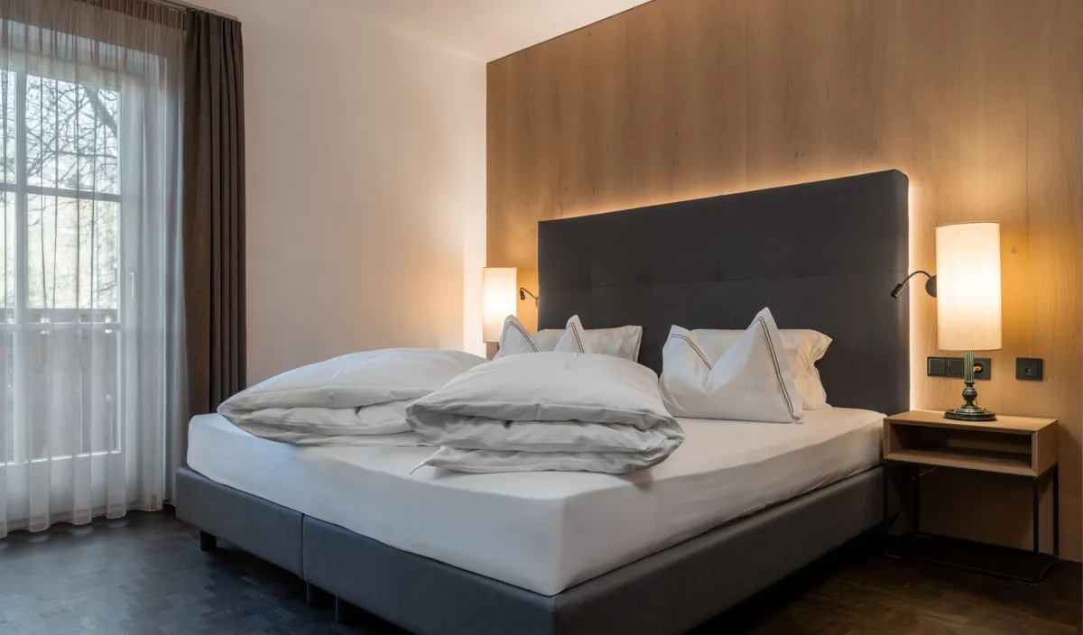 Doppelbett mit Holzwandverkleidung, Nachttischleuchten, Balkontür und Blick auf die Bäume im Zimmer Classic.
