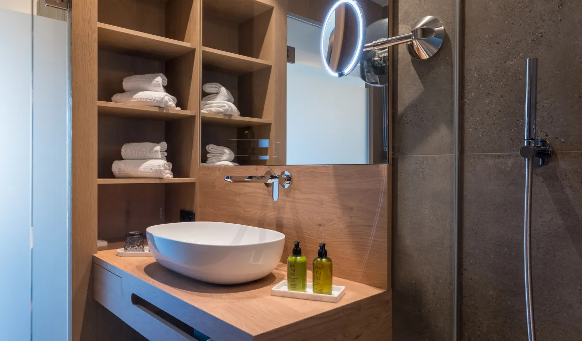 Badezimmerdetail mit Aufsatzwaschbecken, Holzregalen mit Handtüchern und beleuchtetem Kosmetikspiegel.