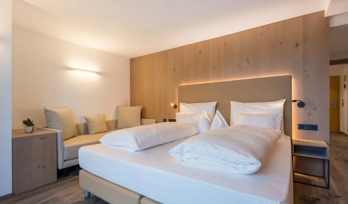 Doppelbett mit gepolstertem Kopfteil, Holzwandverkleidung, Tagesbett und warmem Licht im Zimmer Panorama Superior.