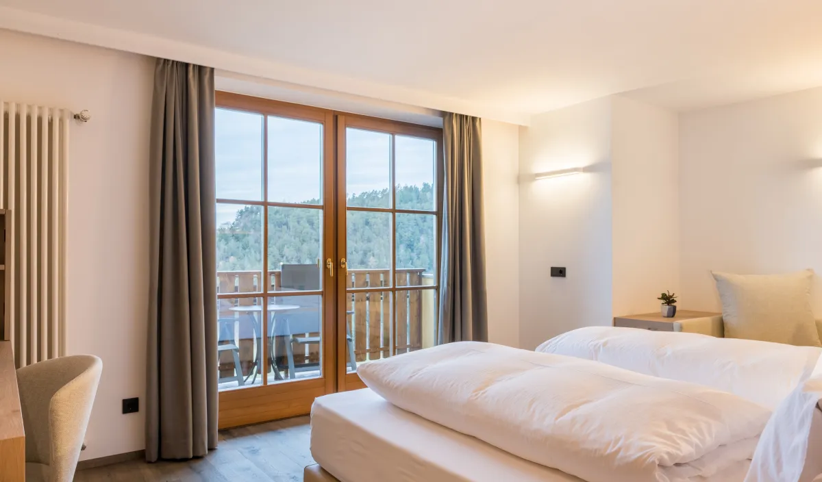 Doppelbett mit Sessel und Balkontür mit Blick auf die bewaldeten Hügel im Zimmer Panorama Superior.