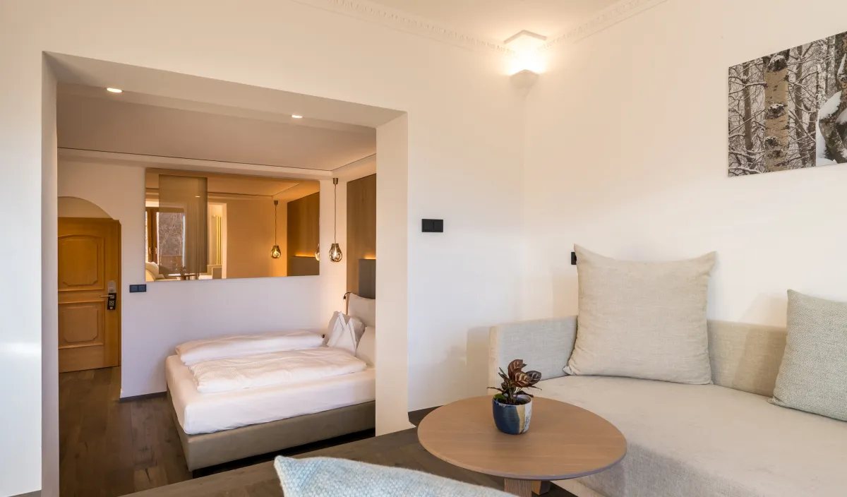 Zona giorno con divano ad angolo, tavolino e vista sulla camera da letto con soffitto a stucco e quadro.