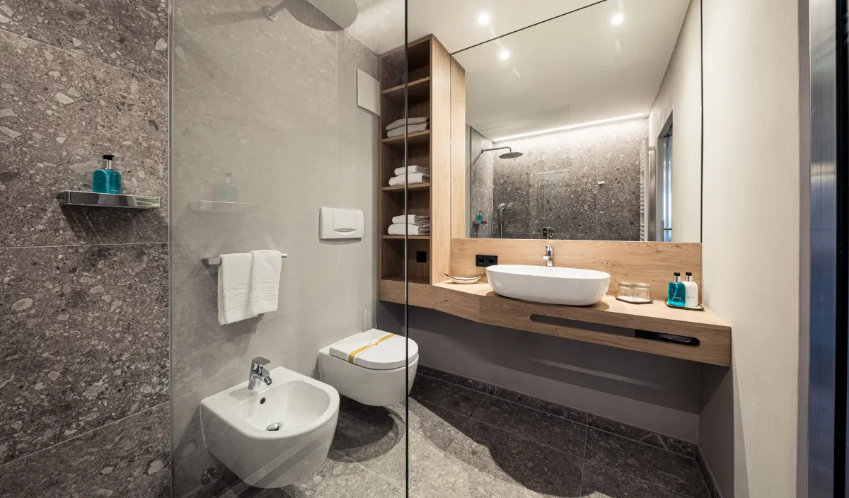 Bagno con doccia in vetro, lavabo d'appoggio su mobile in legno, bidet, WC e piastrelle in terrazzo di pietra naturale.