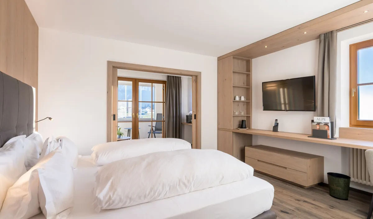 Camera da letto con letto matrimoniale, parete attrezzata in legno con televisore e mensole e passaggio al soggiorno con balcone.