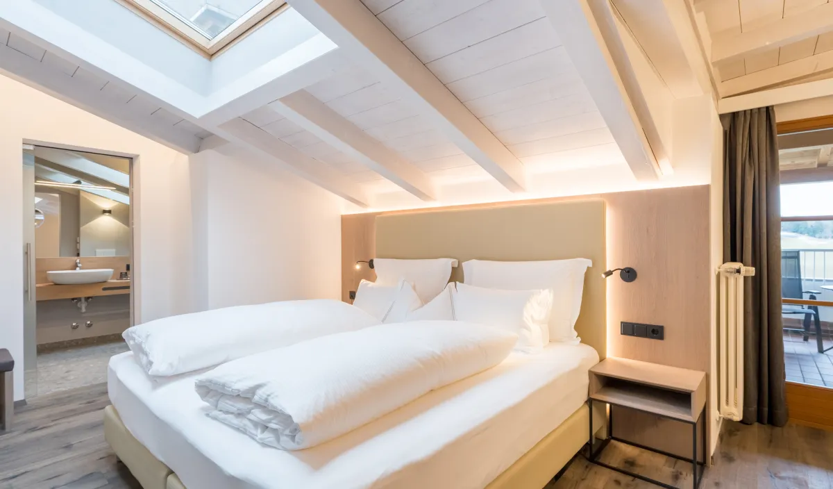 Doppelbett unter der Dachschräge mit Dachfenster, Holzbalkendecke und Blick ins Badezimmer und auf den Balkon.