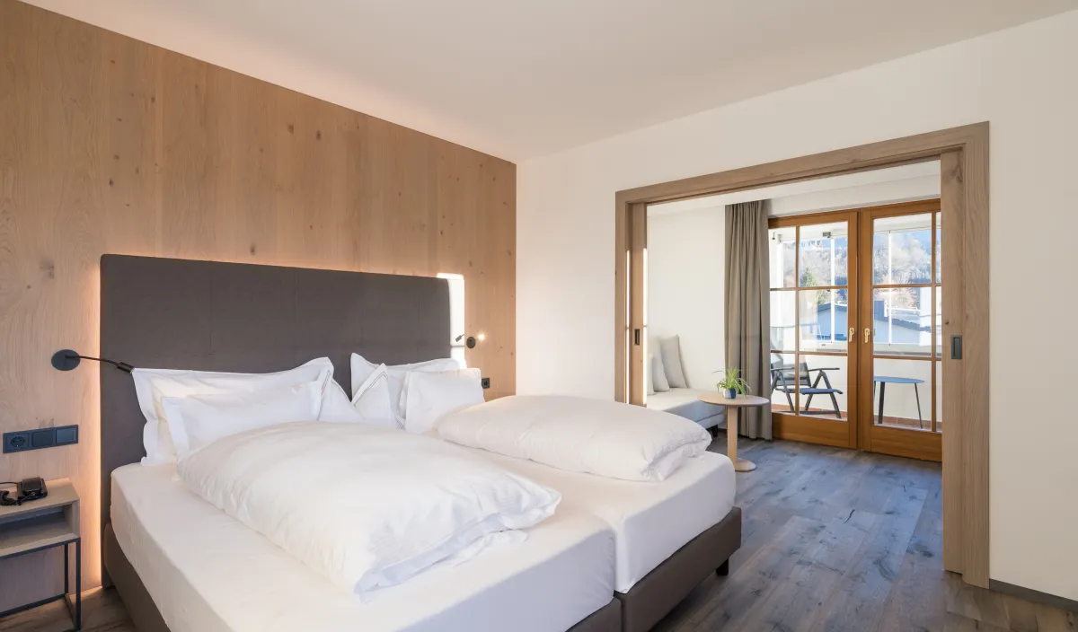 Zona notte con letto matrimoniale, rivestimento in legno e passaggio al soggiorno con divano e porta del balcone.