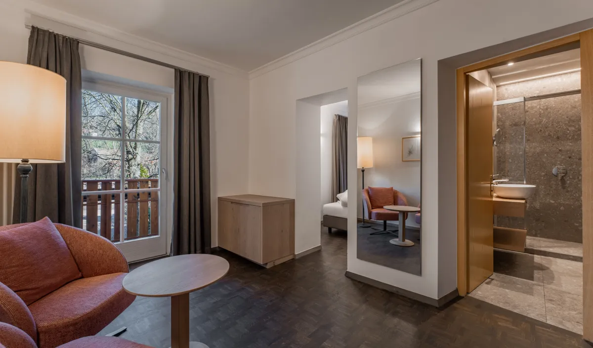 Sitzbereich mit Sessel, Beistelltisch, Stehleuchte, Ganzspiegel, Balkontür und Blick ins Badezimmer im Zimmer Classic.
