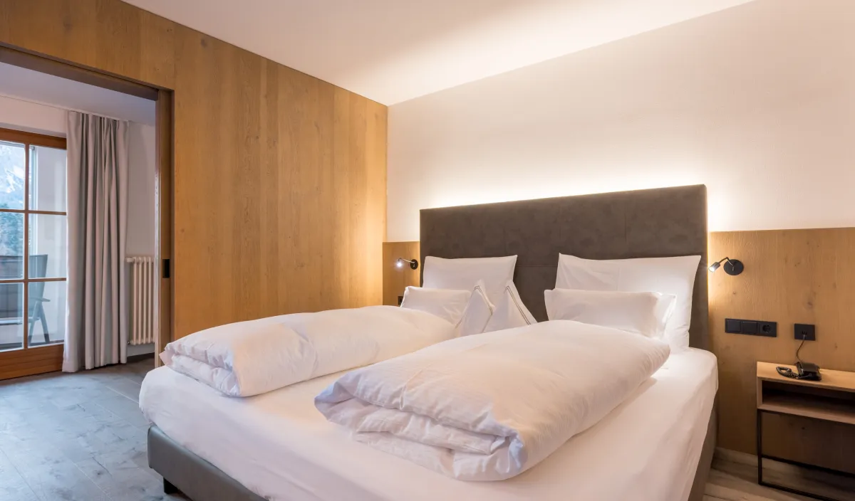 Letto matrimoniale con testiera imbottita, rivestimento in legno e luce calda accanto al passaggio verso il balcone.