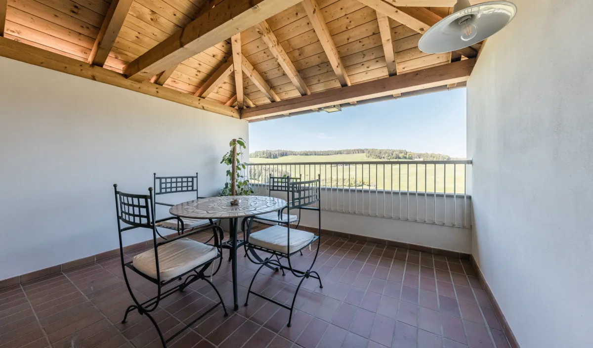 Balcone coperto con tavolo e sedie in ferro battuto, tetto in legno e vista su prati e colline.