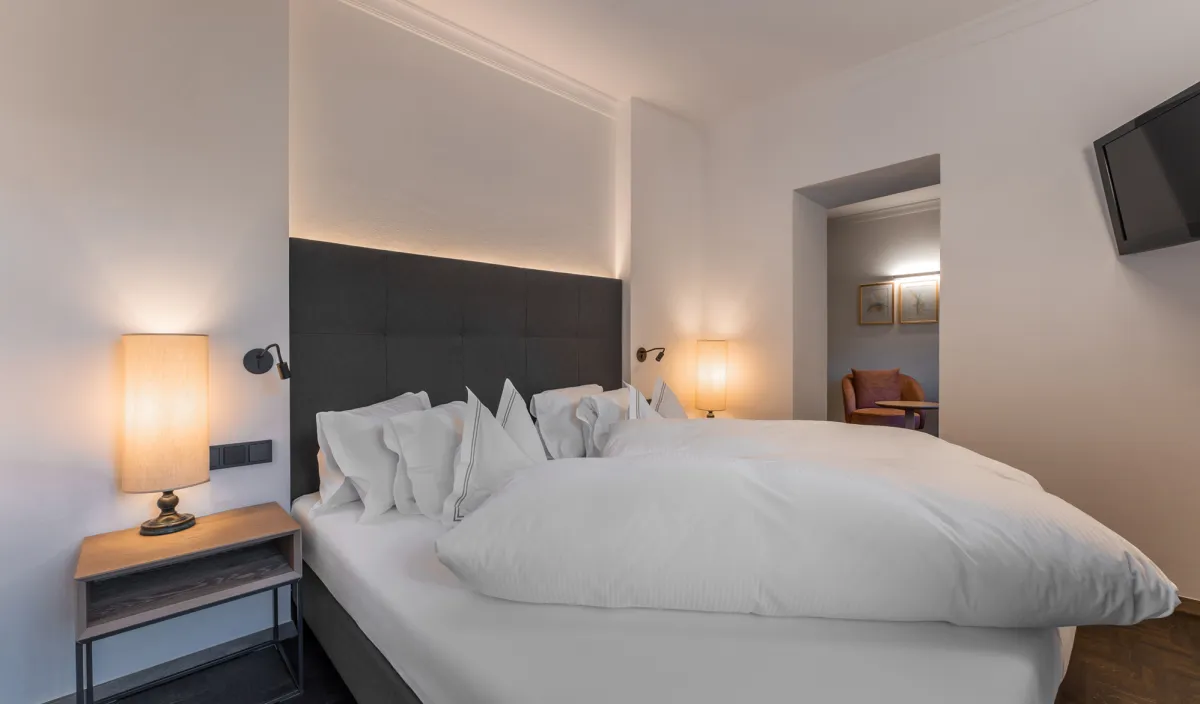 Doppelbett mit beleuchtetem Kopfteil, Nachttischleuchten, Fernseher und Durchgang zum Wohnbereich im Zimmer Classic.