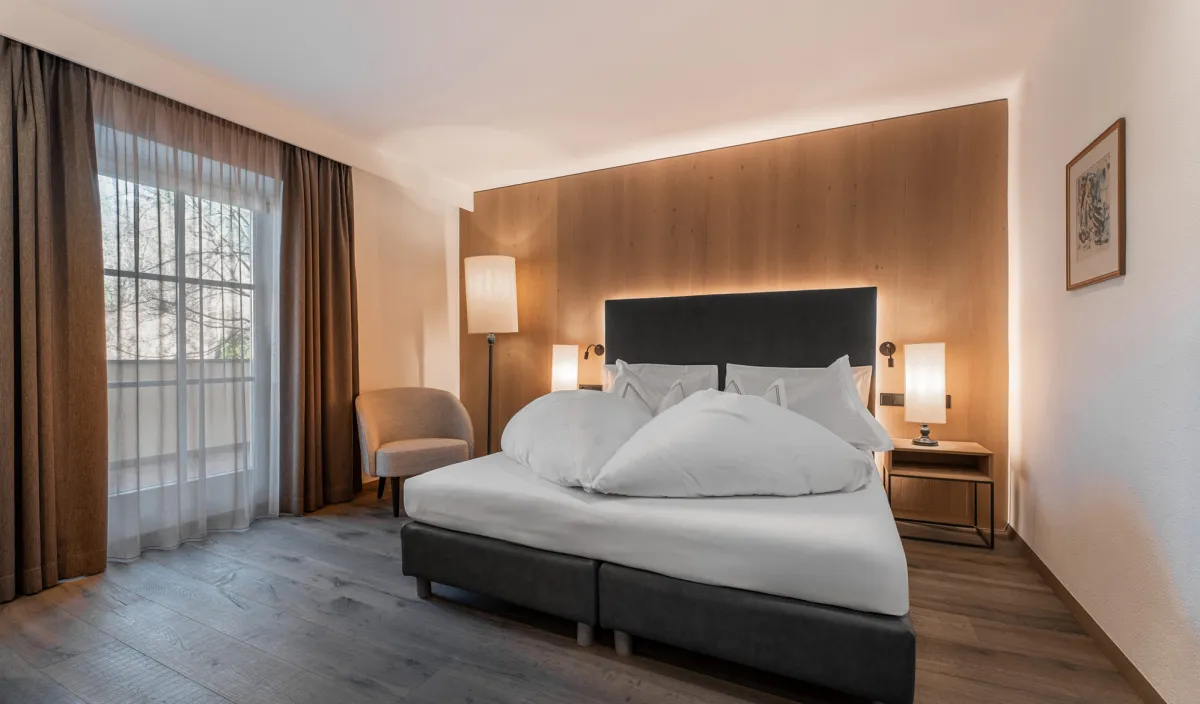 Doppelbett mit Holzwandverkleidung, Nachttischleuchten, Sessel und Fenster mit Blick ins Grüne im Zimmer Classic.