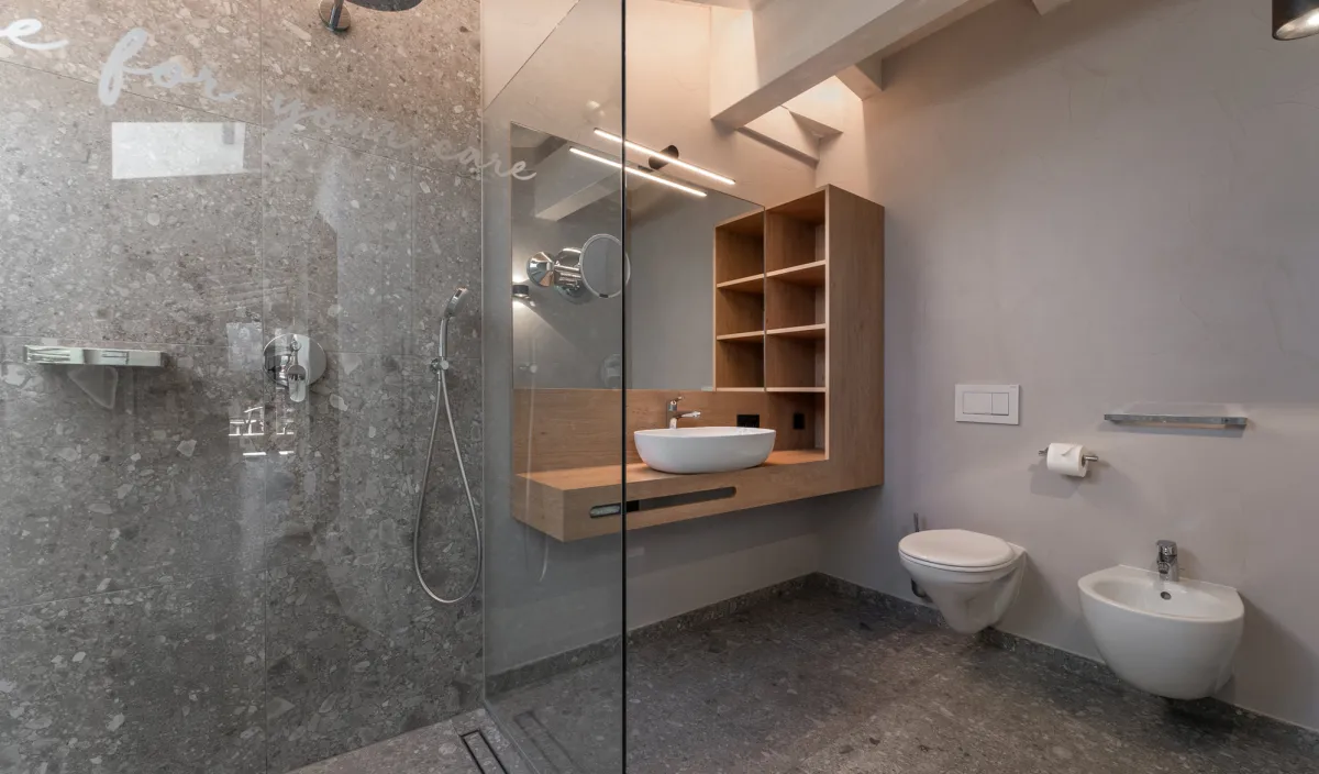 Secondo bagno con doccia in vetro, mobile lavabo sospeso in legno, WC e bidet sotto le travi del tetto.