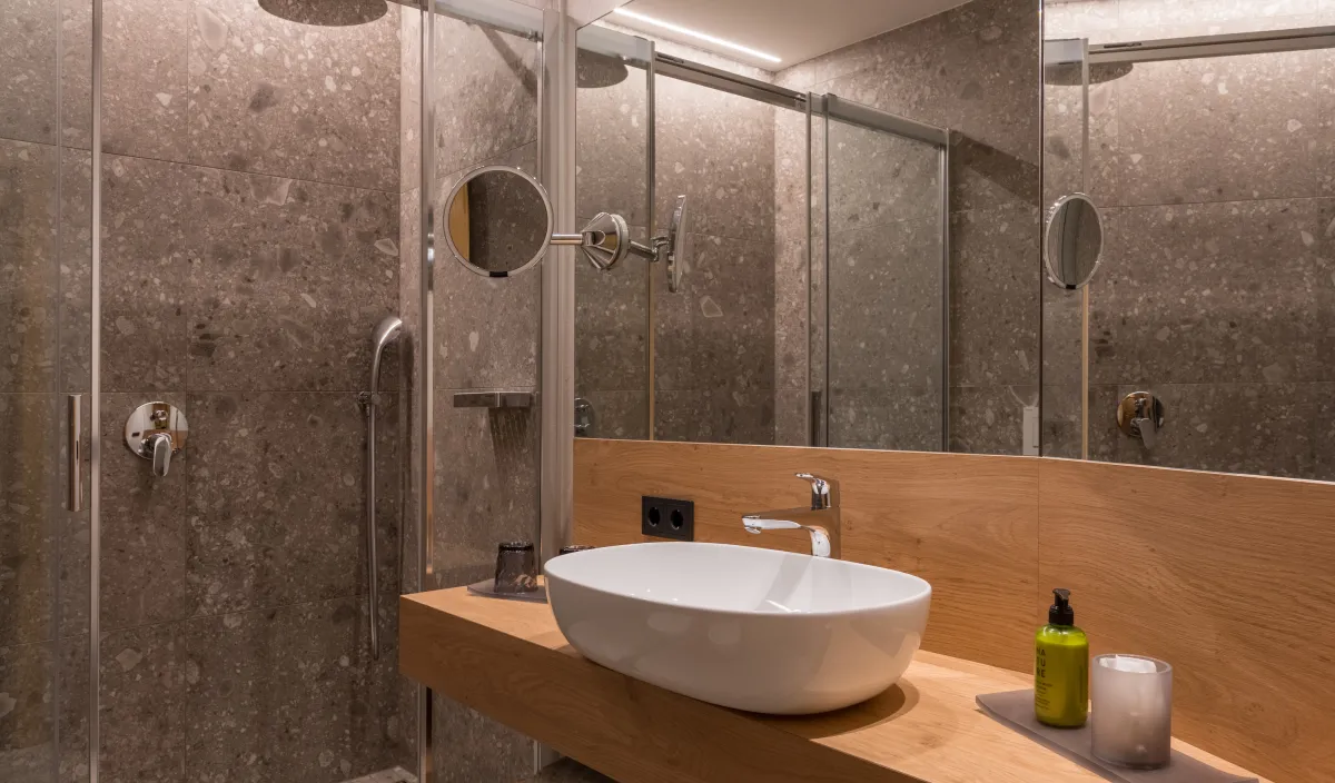 Badezimmerdetail mit Regendusche hinter Glas, Aufsatzwaschbecken auf Holzwaschtisch und Kosmetikspiegel.