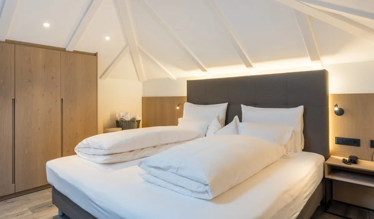 Letto matrimoniale con biancheria bianca, testiera imbottita e armadio in legno sotto il tetto spiovente.