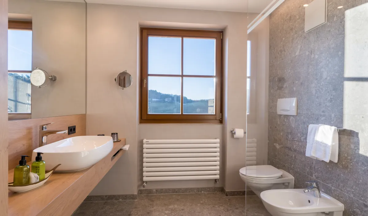 Bagno con lavabo su piano in legno, grande specchio, WC, bidet e finestra con vista sul verde.