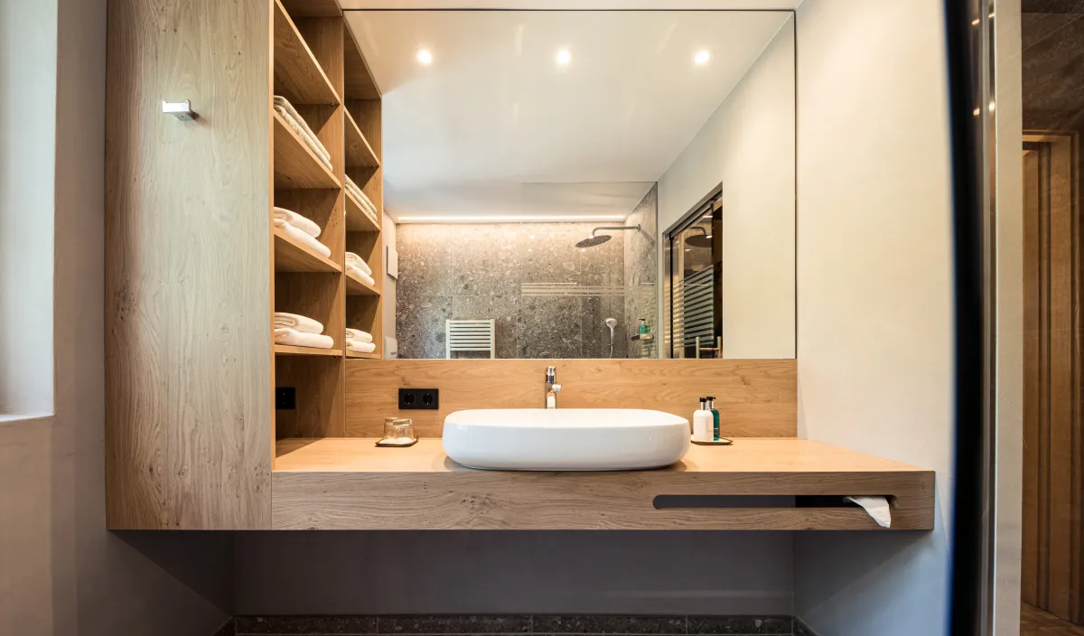 Zona lavabo con bacino d'appoggio su mobile sospeso in legno, mensole per asciugamani e doccia visibile nello specchio.