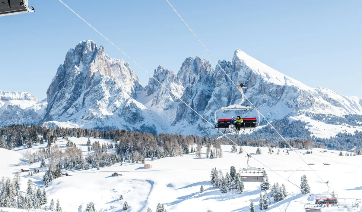 Zwei Personen im Sessellift über der verschneiten Almlandschaft mit den schneebedeckten Dolomiten im Hintergrund