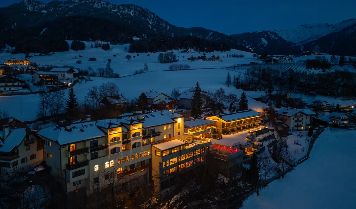 Il Wellnesshotel Alpenflora illuminato in una sera d’inverno a Castelrotto innevata, con vista sulle montagne.