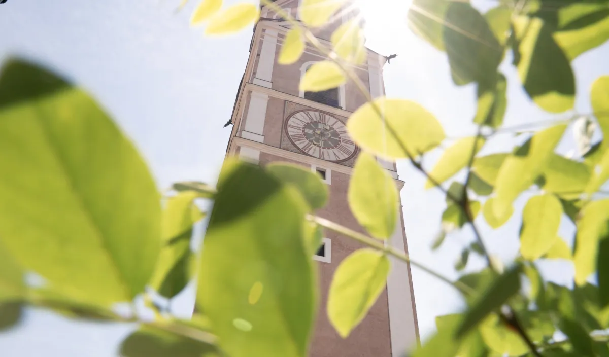 Kirchturm mit Uhr durch grüne Blätter im Sonnenlicht in Kastelruth