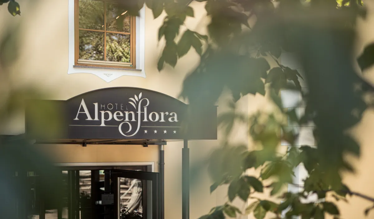 Eingangsschild des Hotels Alpenflora durch grünes Laub am Dorfeingang von Kastelruth.