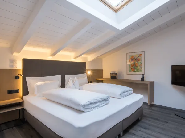 Schlafbereich der Attico Suite mit Doppelbett, weisser Holzbalkendecke, Dachfenster und Blumengemälde.