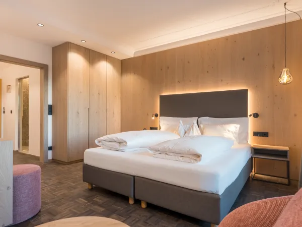 Doppelzimmer Panorama mit Doppelbett, Holzwandverkleidung, rosa Sitzpouf, Holzschrank und Pendelleuchte.