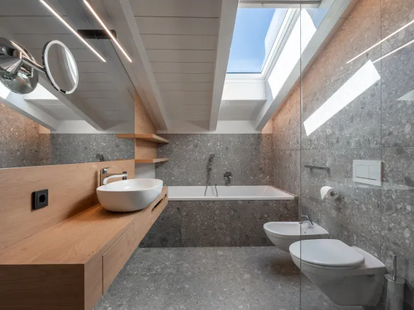 Badezimmer mit Aufsatzwaschbecken auf Holzwaschtisch, Badewanne, Bidet, WC und Dachfenster mit blauem Himmel.