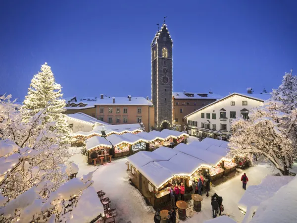 Verschneiter Weihnachtsmarkt mit beleuchtetem Baum und Stadtturm bei Nacht in Südtirol