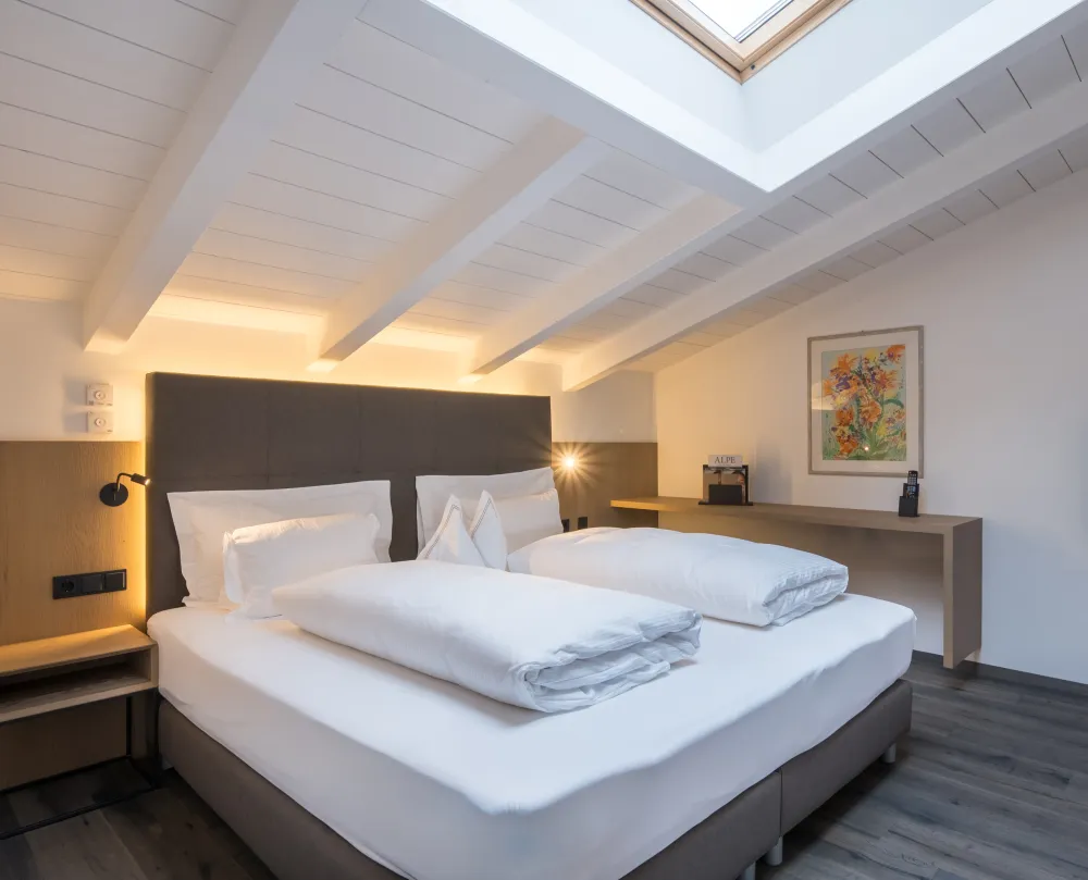 Schlafbereich der Attico Suite im Wellnesshotel Alpenflora mit Doppelbett, Dachfenster und weißer Holzbalkendecke.