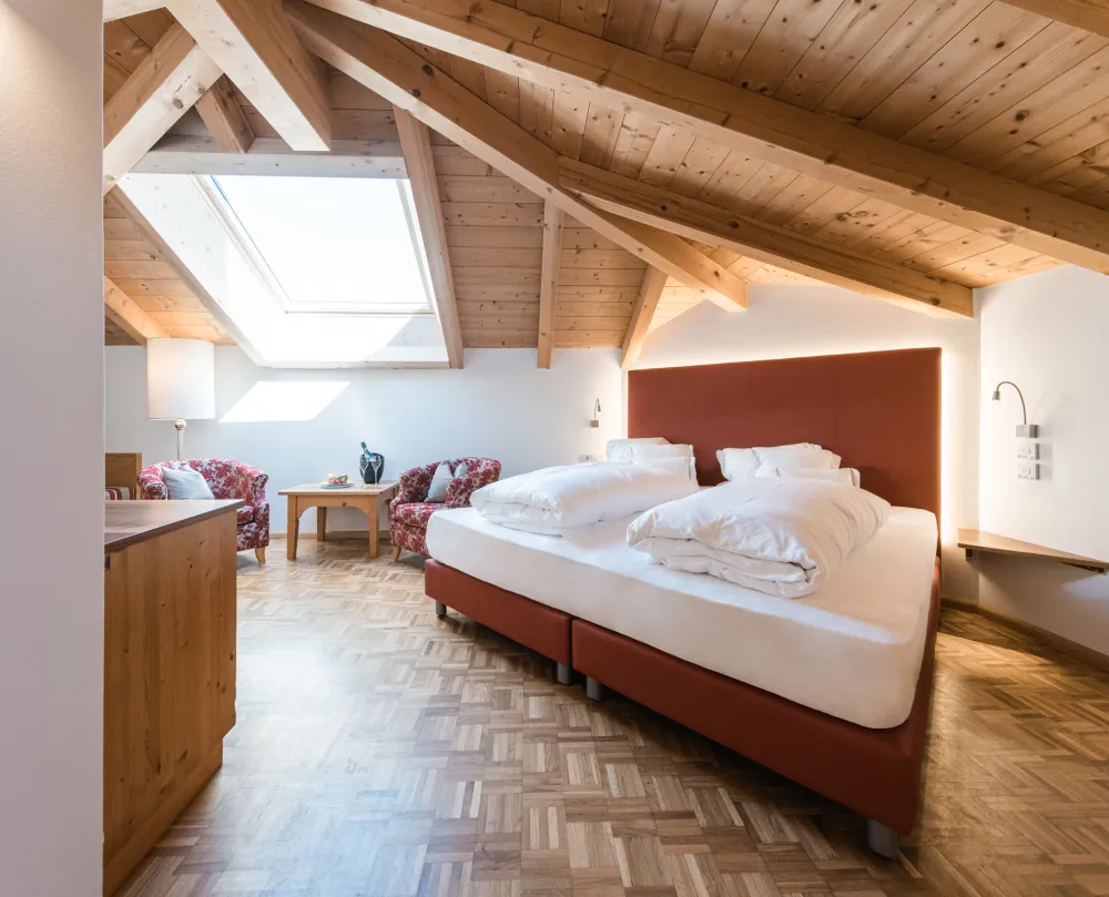 Doppelzimmer Attico im Wellnesshotel Alpenflora mit Doppelbett, Dachfenster, Holzbalkendecke und zwei gemusterten Sesseln.