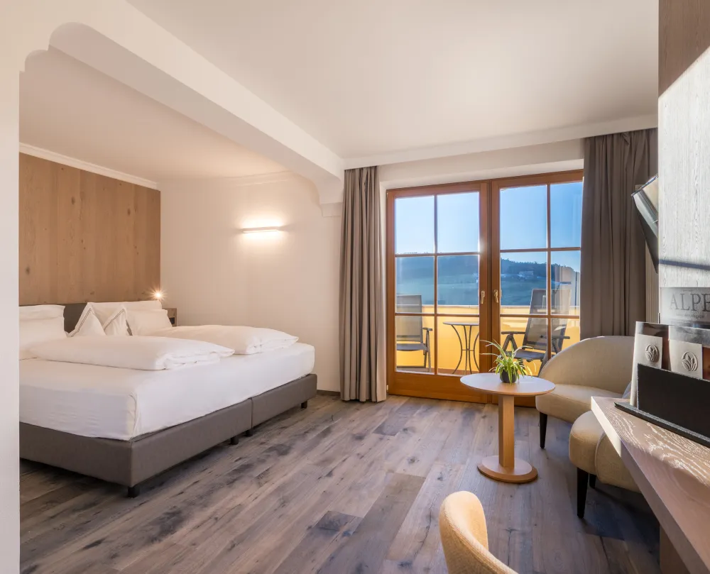 Doppelzimmer Panorama Superior im Wellnesshotel Alpenflora mit Doppelbett, Sessel und Balkontür mit Blick ins Grüne.