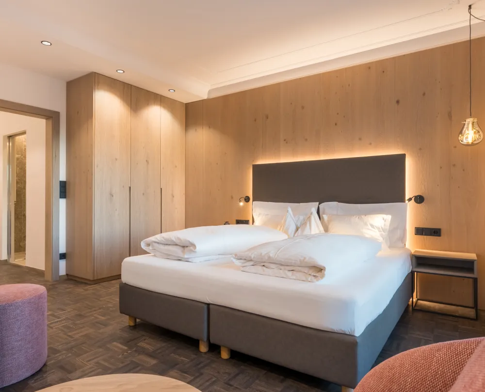 Doppelzimmer Panorama im Wellnesshotel Alpenflora mit Doppelbett, Holzschrank, Holzwandverkleidung und rosa Sitzpouf.