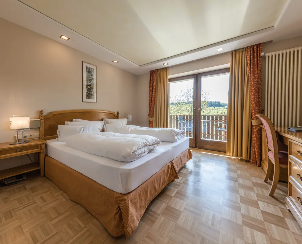 Doppelzimmer Sun im Wellnesshotel Alpenflora mit Doppelbett, Nachttisch, Schreibtisch und Balkontür mit Blick ins Grüne.